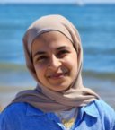 A young woman wearing a beige hijab and a blue button-down shirt smiles at the camera in front of the beach.