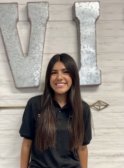 A young latina woman with long dark brown hair wearing a black scrub top smiles at the camera in front of a white wood panel background.