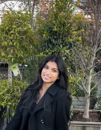 A young indian woman with long dark brown hair wearing a black trench coat leans to her right and smiles at the camera in front of a leafy green plant.