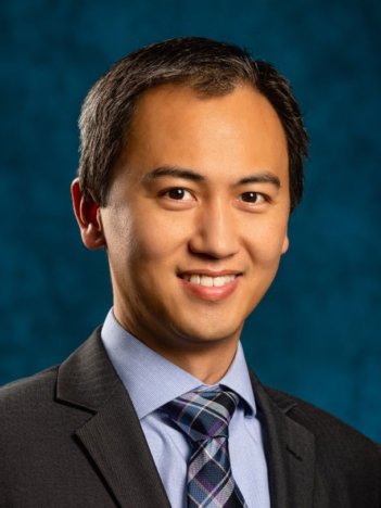 An asian man with short dark brown hair wears a blue collared shirt, plaid blue tie, and dark grey suit jacket and smiles at the camera in front of a navy background.