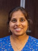 An indian woman with brown hair pinned back into a bun wears a florally patterned blue blouse and smiles at the camera. 