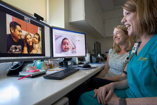 Two people look at two monitors. One with two adults and a child and another monitor with a baby
