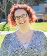 A person with curly red hair and glasses smiles at the camera outdoors. They are wearing a black and white patterned shirt.