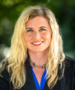 A person with long blonde hair, smiling and looking at the camera. She is wearing a black blazer over a blue shirt and a beaded necklace.