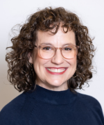 Smiling person with curly dark hair and glasses wearing a dark turtleneck against a plain light background.