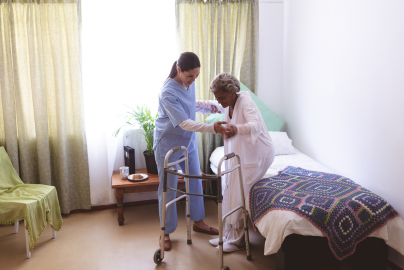 A caregiver assists a person in a white gown stand up from a bed, adjusting a walker.