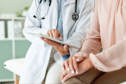 A doctor in a white coat with a stethoscope around their neck is showing something on a tablet to a patient sitting down.