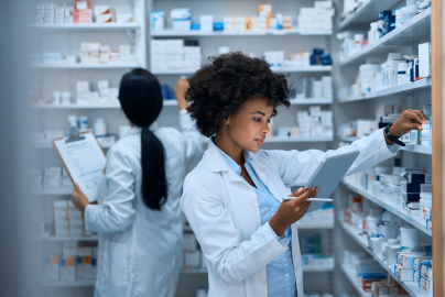 A pharmacist holds a tablet and reaches for a product on a shelf, while another person stands behind them.