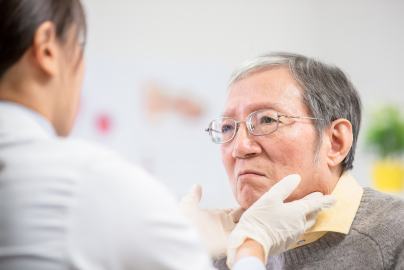 A healthcare worker in white scrubs examines an elderly patient wearing glasses and a grey sweater.