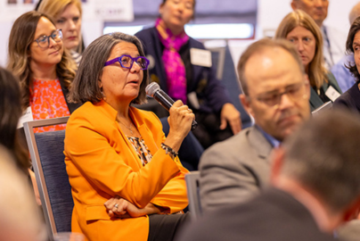 A woman in an orange coat speaks into a microphone while seated in a crowd.