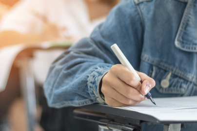 The right arm of a student in a jean jacket writes on a desk in a classroom.