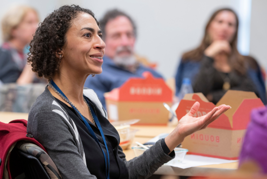 A person wearing a grey coat over a black shirt and curly hair speaks while seated next to a table..