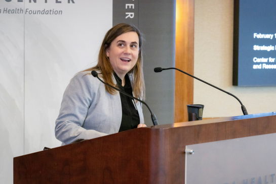 A person wearing a grey coat with shoulder length hairs speaks behind a podium.