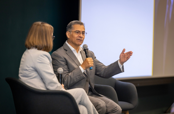 A seated person in a suit speaks into a microphone next to another person seated and wearing in a suit.