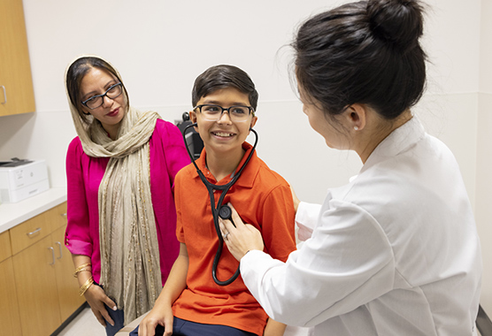 Primary care provider using stethoscope to have smiling boy hear his heart while mother stands nearby.