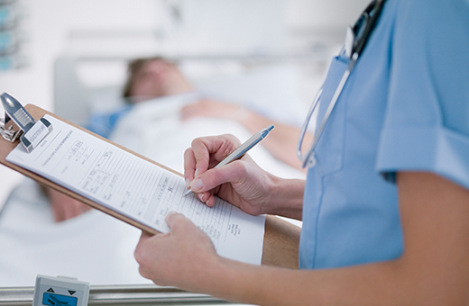 A nurse writes on a clipboard while a man lies in a hospital bed behind her.