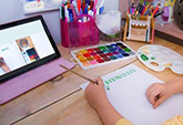 A child’s hands seen painting onto paper with iPad next to them. 