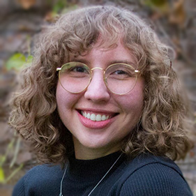 Megan Tudor, with curly brown hair and glasses, smiles for the camera. 
