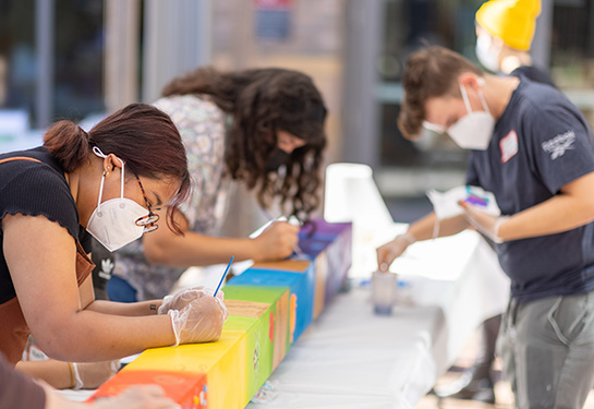 Two young women and one young man are leaning over a colorful pole that’s laying on a table, painting designs and words on to it. 