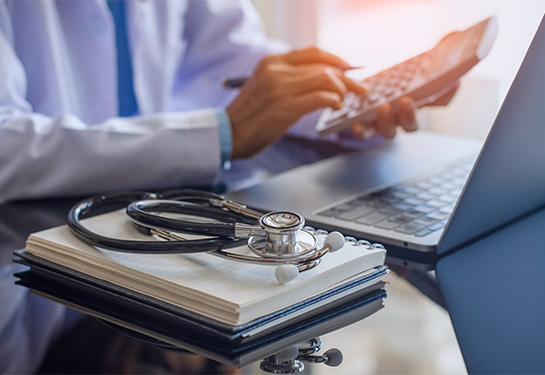 Person in white doctor’s coat sits next to a stethoscope resting on a notebook