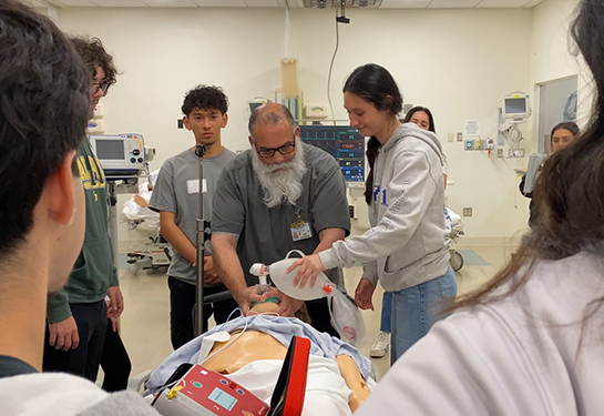 A man with a beard stands between high school students and teaches them to pump air into a training manikin’s nose and mouth 