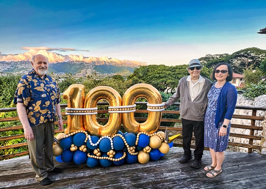 From left, Mark Mannis, Ernest E. Tschannen and Michele Lim at the birthday celebration, stand next to a metallic balloon in shape of 100.