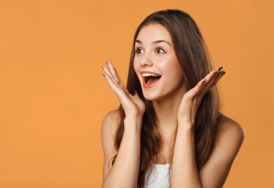 Surprised, happy woman with long hair smiles with open hands near her cheeks. 
