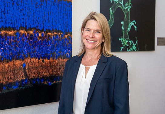 Marie Burns, wearing a blue suit and white shirt, stands next to artwork for a portrait.