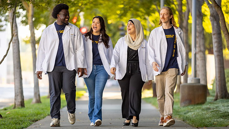 Four diverse medical students in white coats walk together on a tree-lined campus path, laughing and chatting cheerfully.