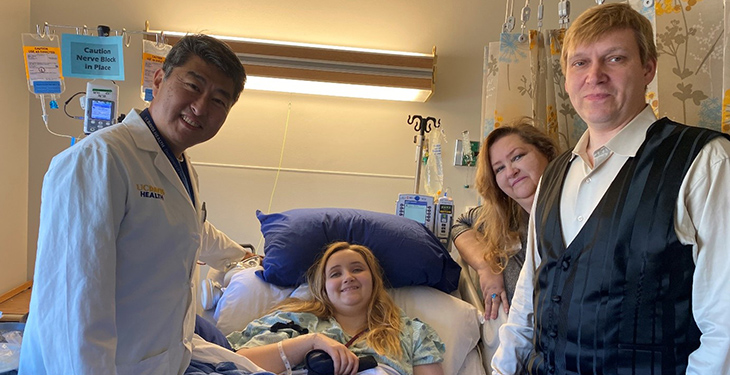 In the center, Tiffany Balts in a hospital bed after her surgery. Neurosurgeon Kee Kim standing to Tiffany’s right and her mother and dad to her left.