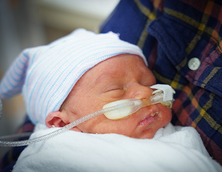 An infant with a nasal oxygen tube sleeps in a person’s arms while wearing a white and blue stocking cap.