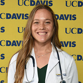 Kelly Weldon, wearing a white coat, stands in front of a backdrop that says UC Davis Health in blue and yellow.