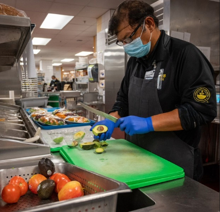 A food services employee prepares fresh avocado in a commercial kitchen, with fruits, vegetables and meal ingredients arranged nearby for food preparation.