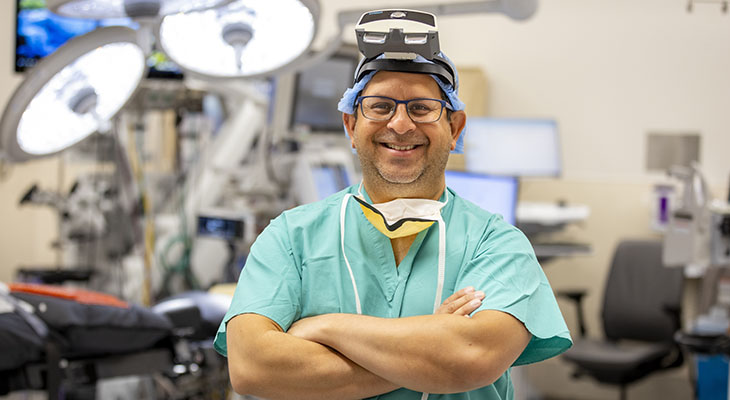 Surgeon Safdar Khan, in light green scrubs, stands in an operating room with augmented reality camera strapped to his head.