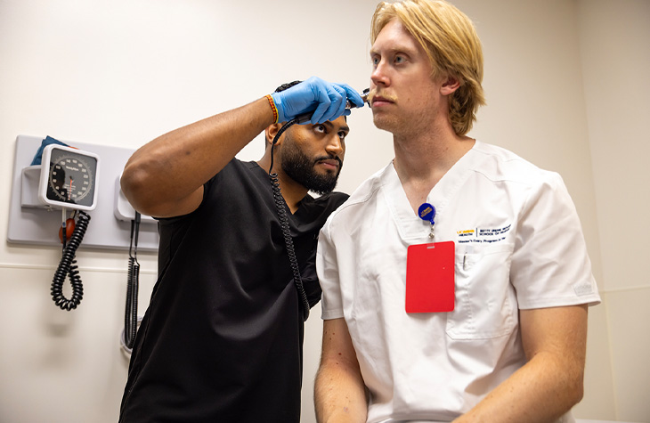 Male nursing student wearing black scrubs and blue gloves practices an eye examination on another male student in white scrubs.