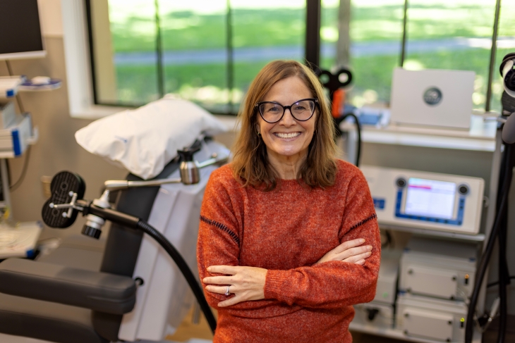 A smiling woman wearing glasses and an orange sweater stands in a room with medical equipment. 