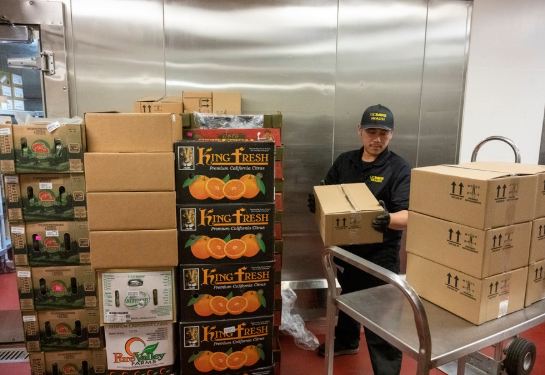 Person moves boxes of food supplies from a delivery cart inside a storage area lined with stacked produce and shipping cartons.