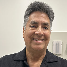 A man with black hair, wearing a black shirt, poses for a photo in a doctor's office