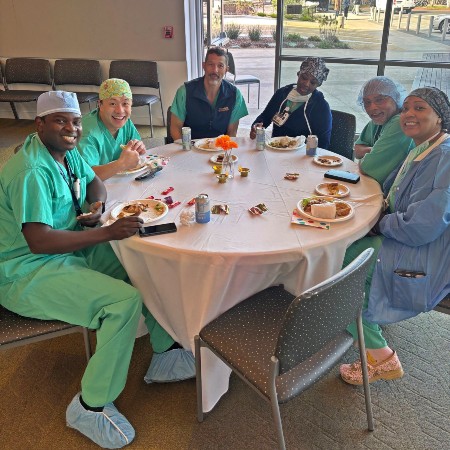 Staff wearing scrubs are sitting at a table, eating food and smiling