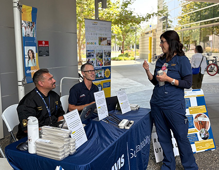 Two adults sit behind a table containing reusable utensil kits while another adult talks with them. 