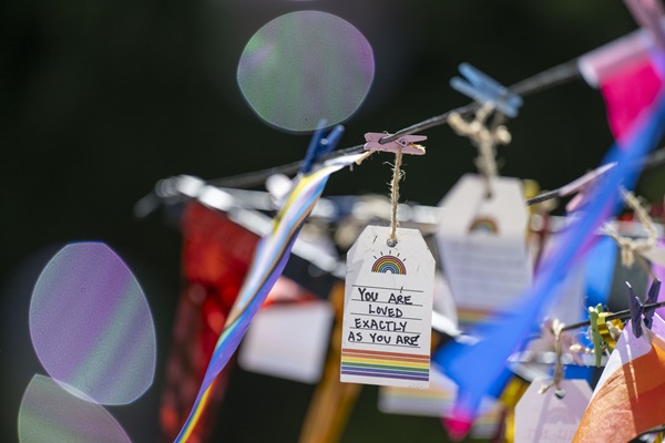 Handwritten Pride messages hang from strings with rainbow ribbons and decorations outdoors.