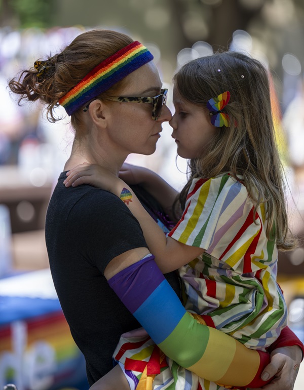 Adult holding a child, both wearing rainbow-colored clothing and accessories at a Pride event.