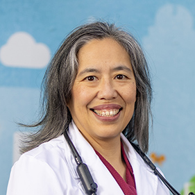 A woman with long gray-black hair, wearing a white doctor's coat, smiles for the camera.