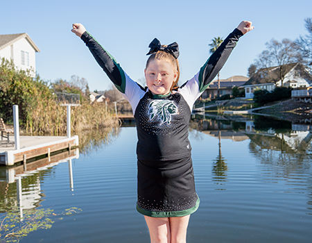 A young girl stands on a deck with water behind her. She wears a cheerleader uniform and stands with arms in a “Y” shape.
