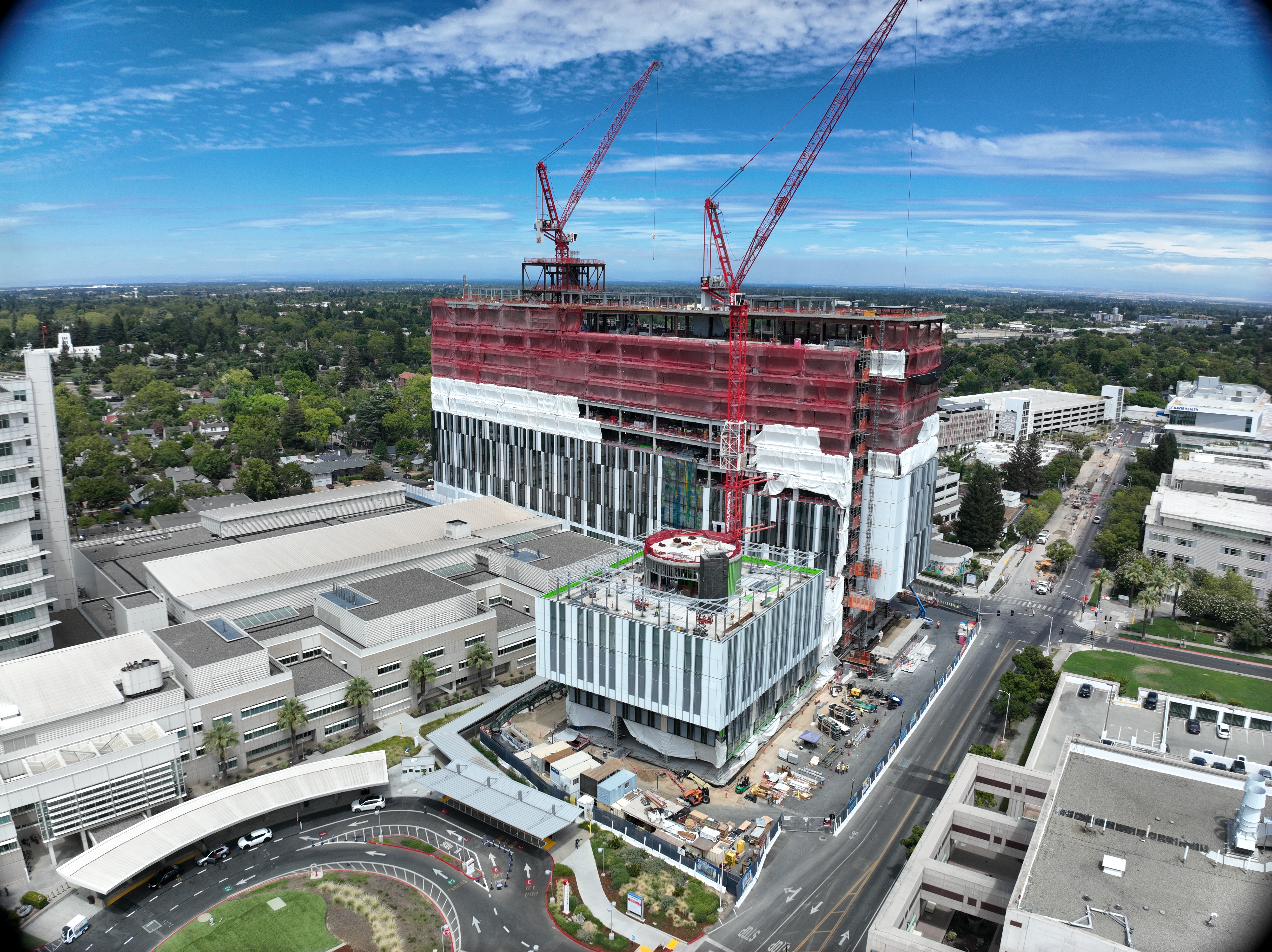 Aerial view of California Tower under construction with red safety netting, white exterior wrap and two cranes above the structure on the Sacramento campus.