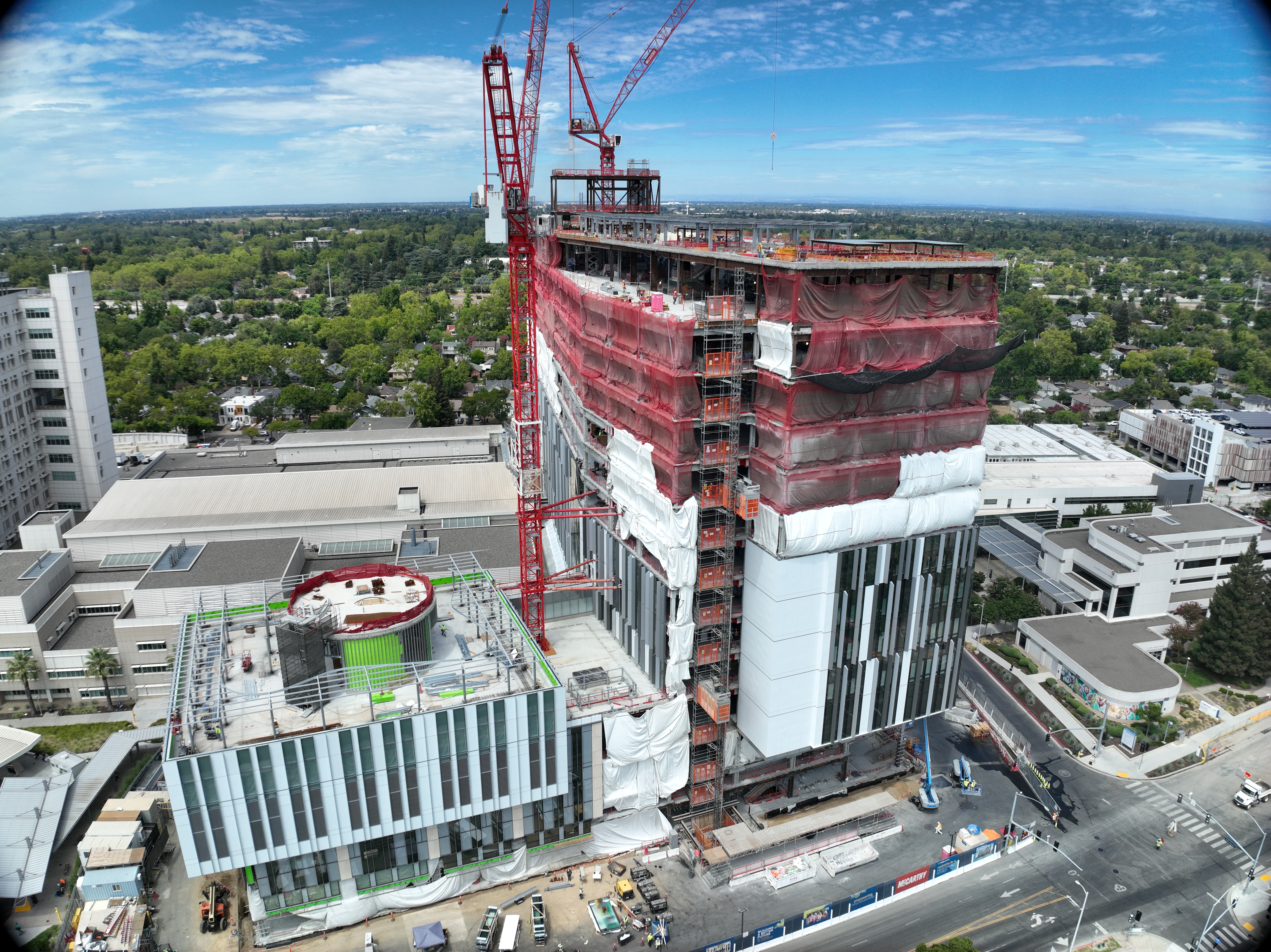 Aerial view of California Tower under construction with red safety netting, white exterior wrap and two cranes above the structure on the Sacramento campus.