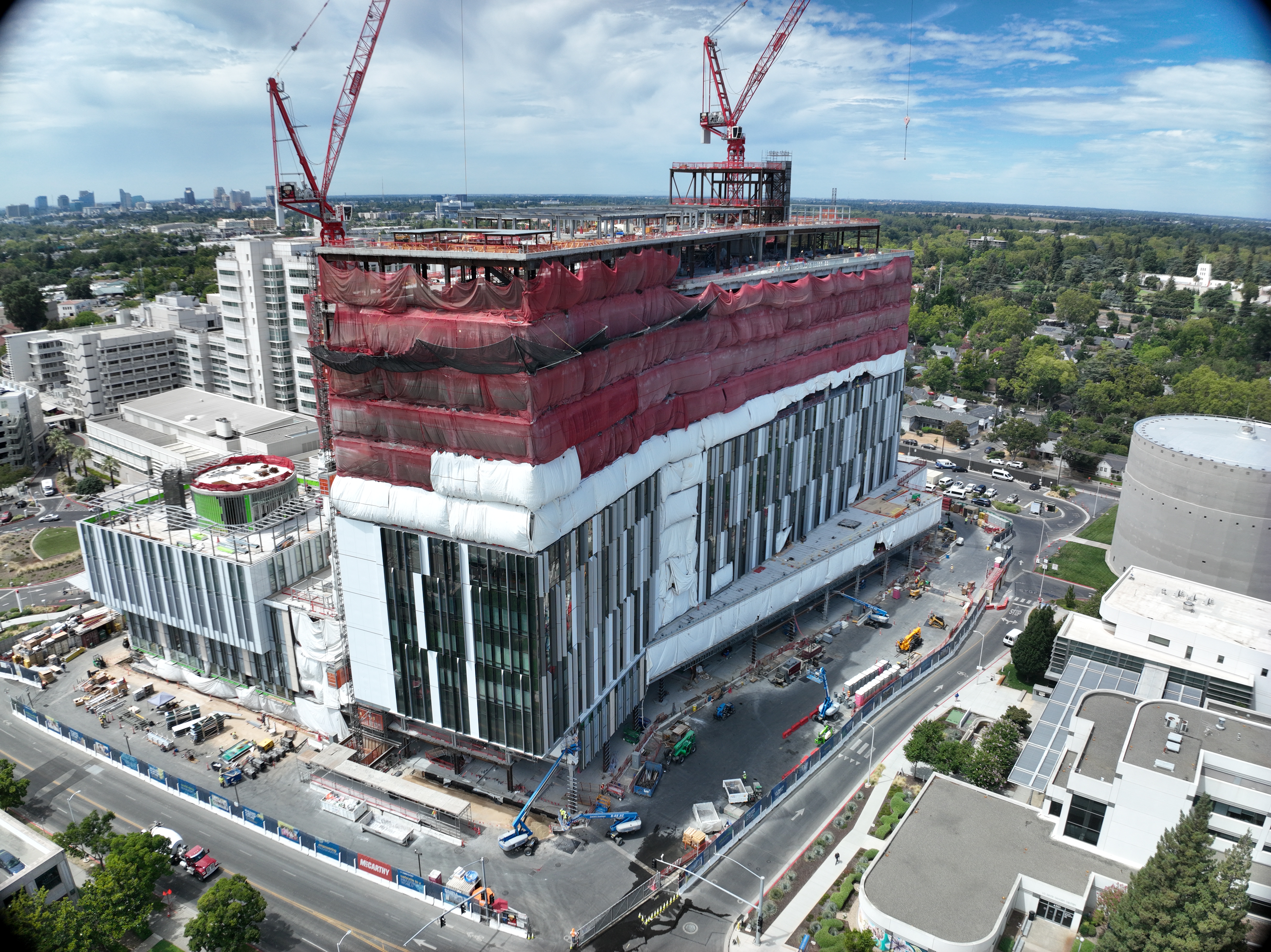 Aerial view of California Tower under construction with red safety netting, white exterior wrap and two cranes above the structure on the Sacramento campus.