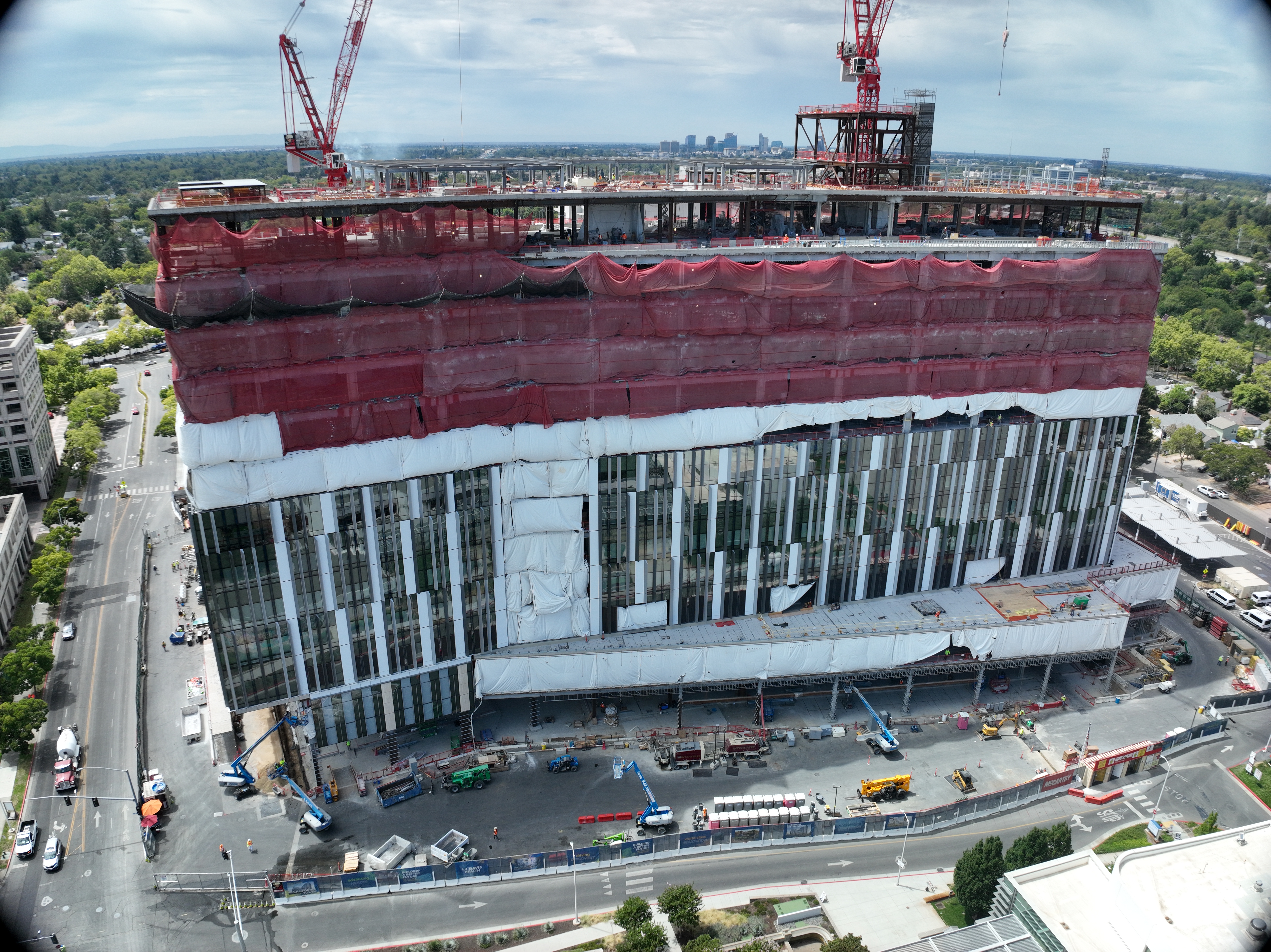 Aerial view of California Tower under construction with red safety netting, white exterior wrap and two cranes above the structure on the Sacramento campus.