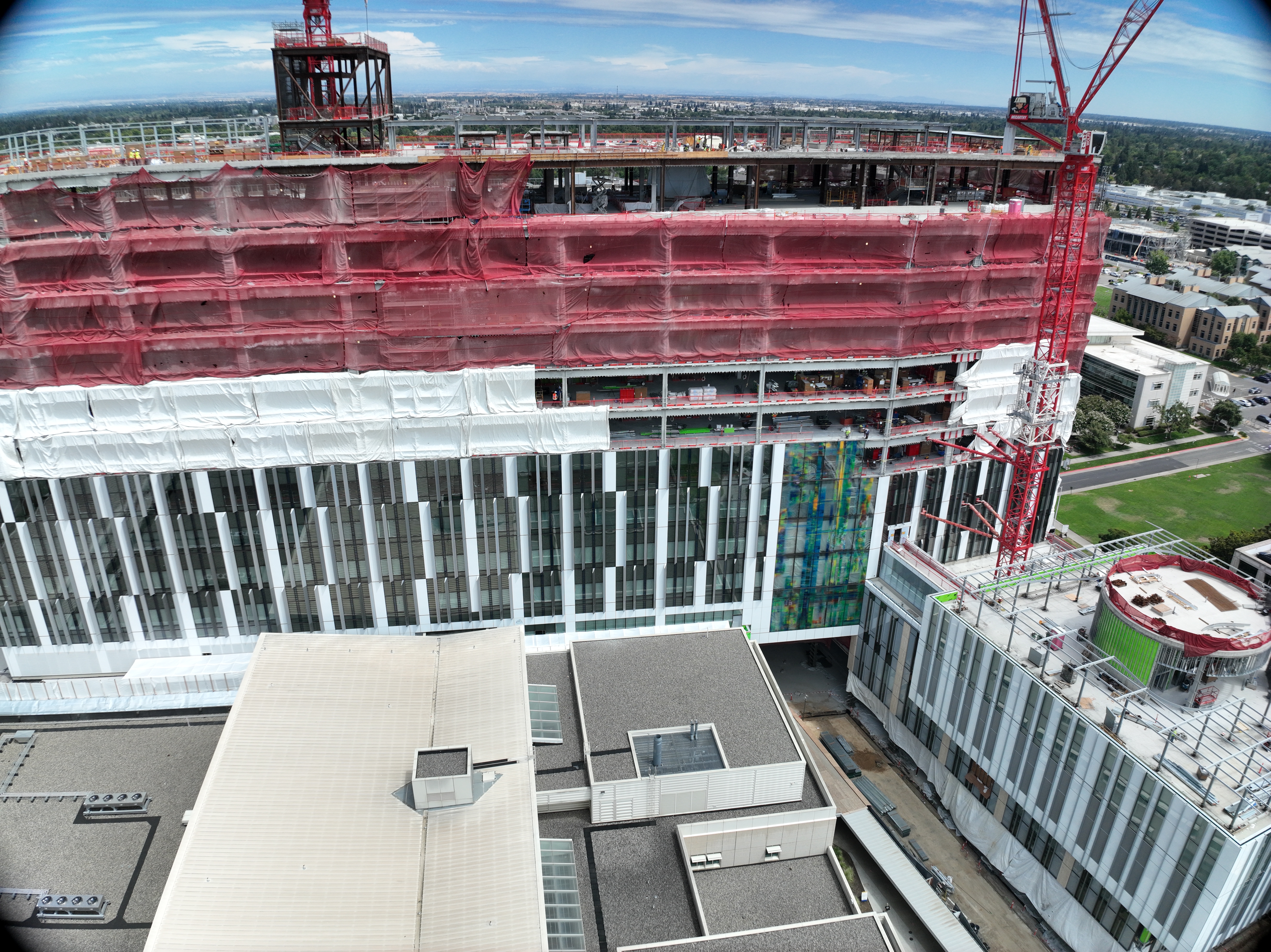Aerial view of California Tower under construction with red safety netting, white exterior wrap and two cranes above the structure on the Sacramento campus.