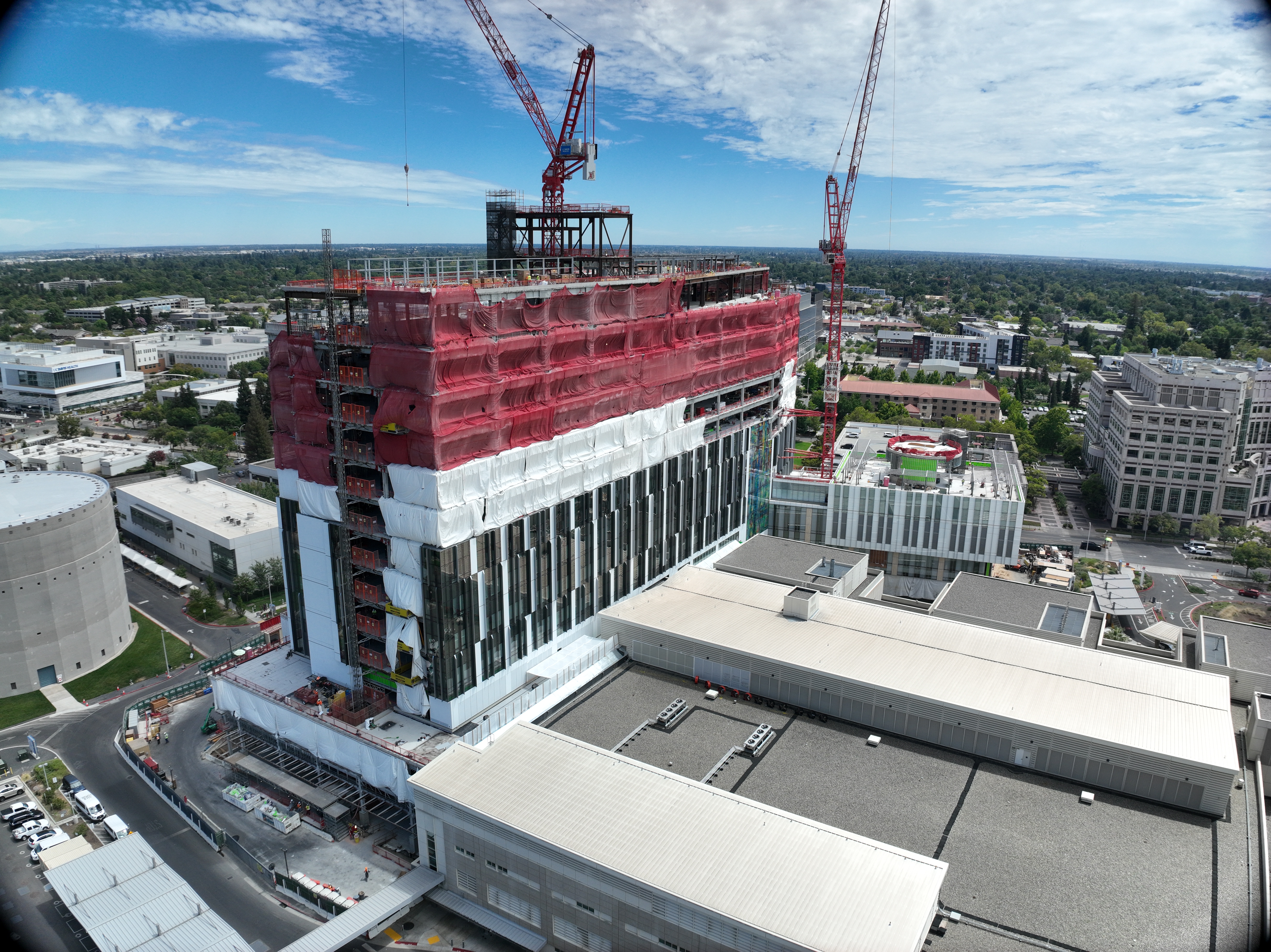 Aerial view of California Tower under construction with red safety netting, white exterior wrap and two cranes above the structure on the Sacramento campus.